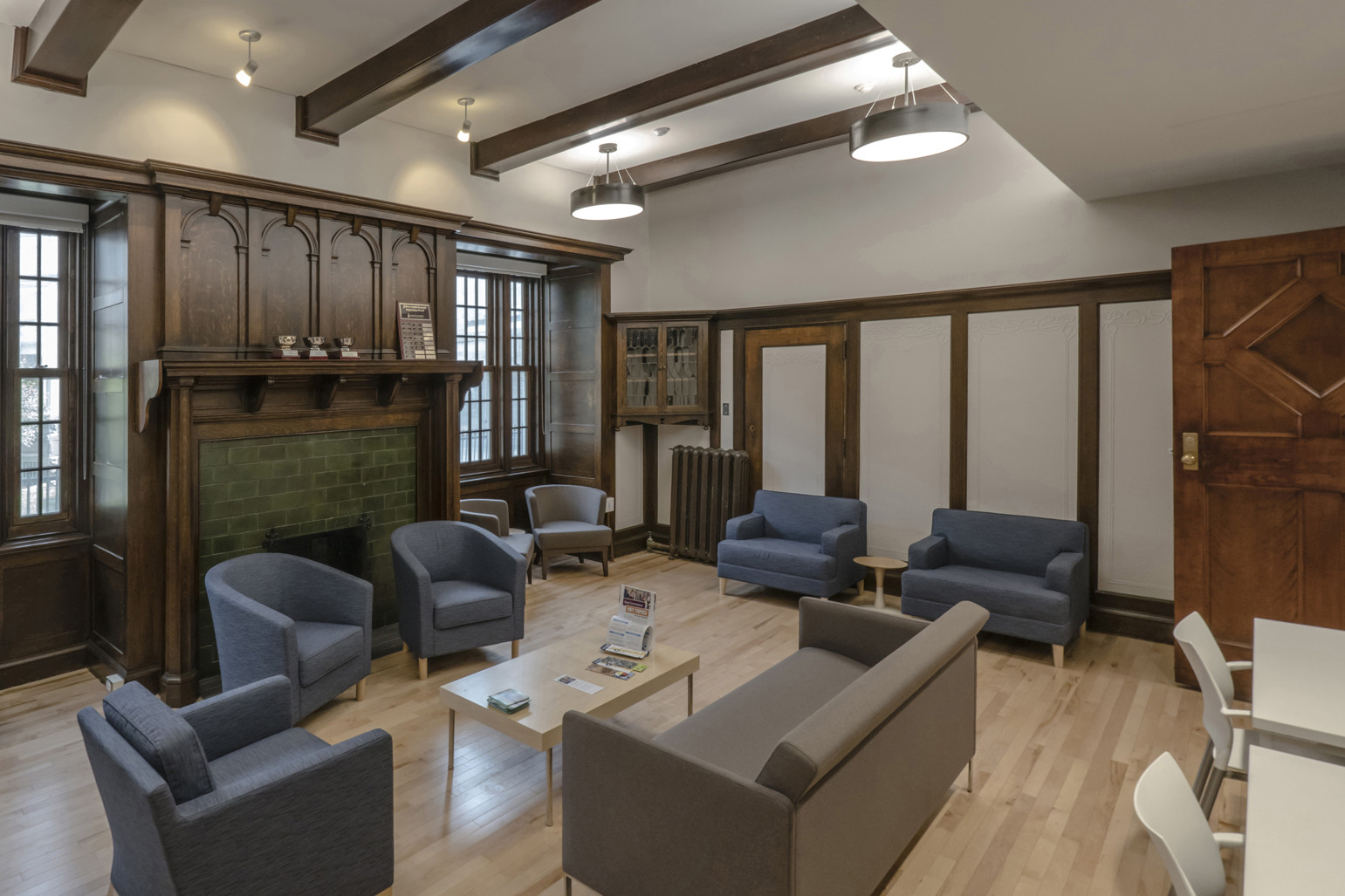 Student lounge with blue armchairs, grey couch, green brick fireplace, wood wall detailing, exposed wood beam ceiling, and large windows