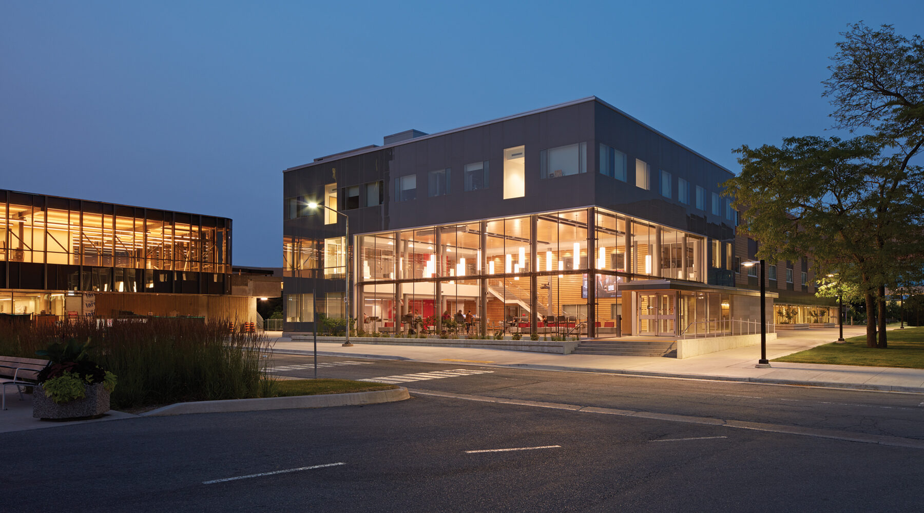 View of building from across the street featuring illuminated double height student commons at dusk