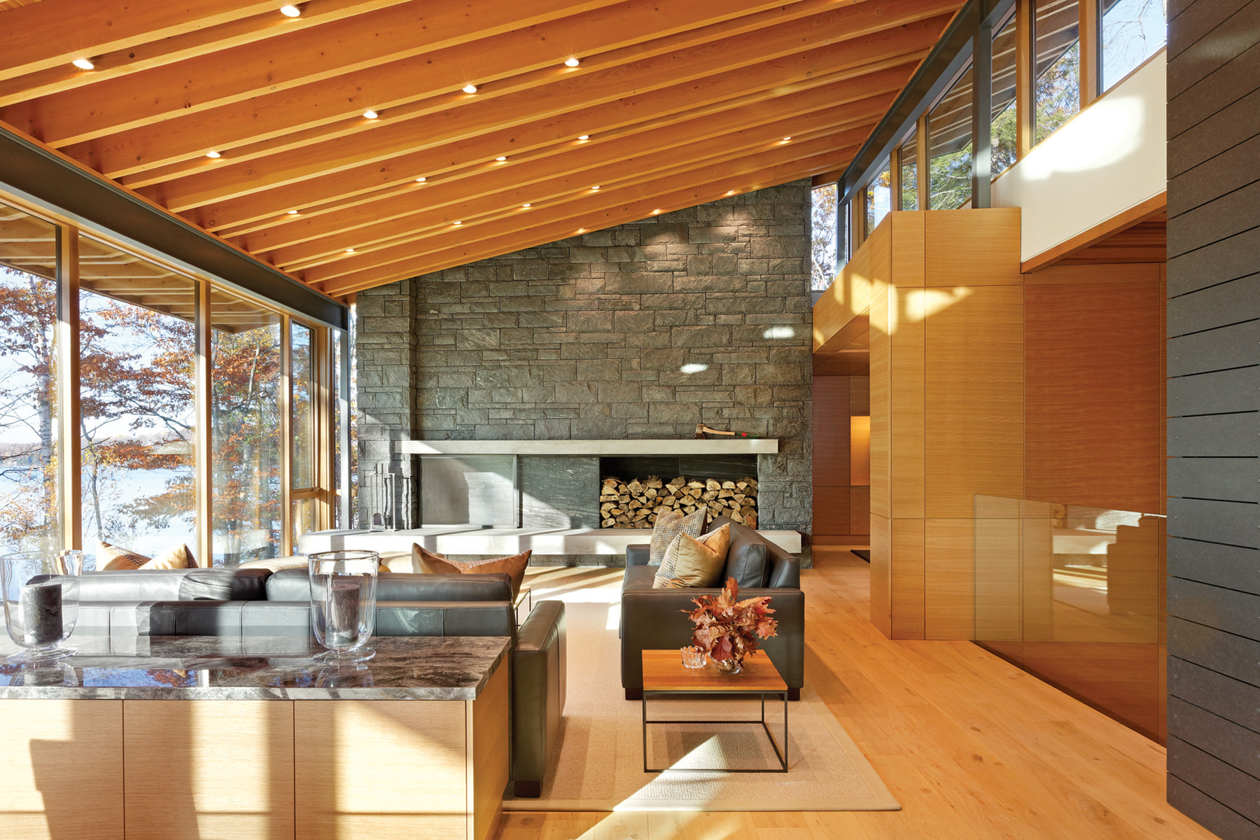 Living room with fireplace, slanted rafter ceiling with clerestory windows on the right and glazed wall on the left with views of lake and trees