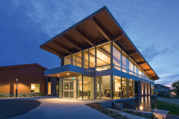 Front entrance to illuminated two-storey building with glass facade and wood cantilever roof at dusk