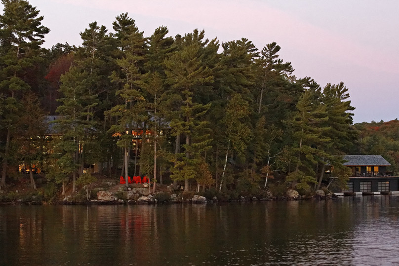 View of illuminated cottage and boathouse from lake mostly obscured by trees at sunset