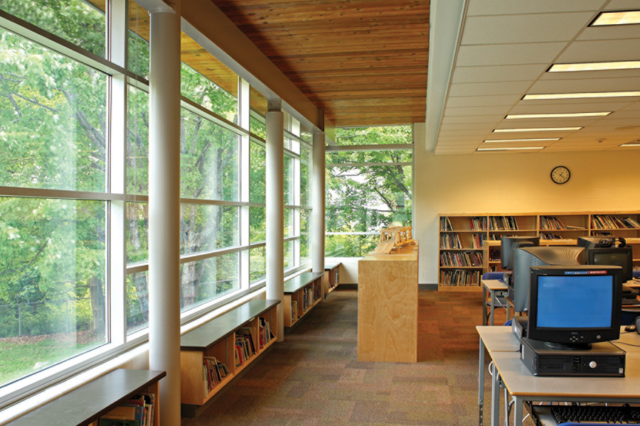Library with bookshelves and computers on right and large steel framed windows, exposed pillars and wood slat ceiling on left