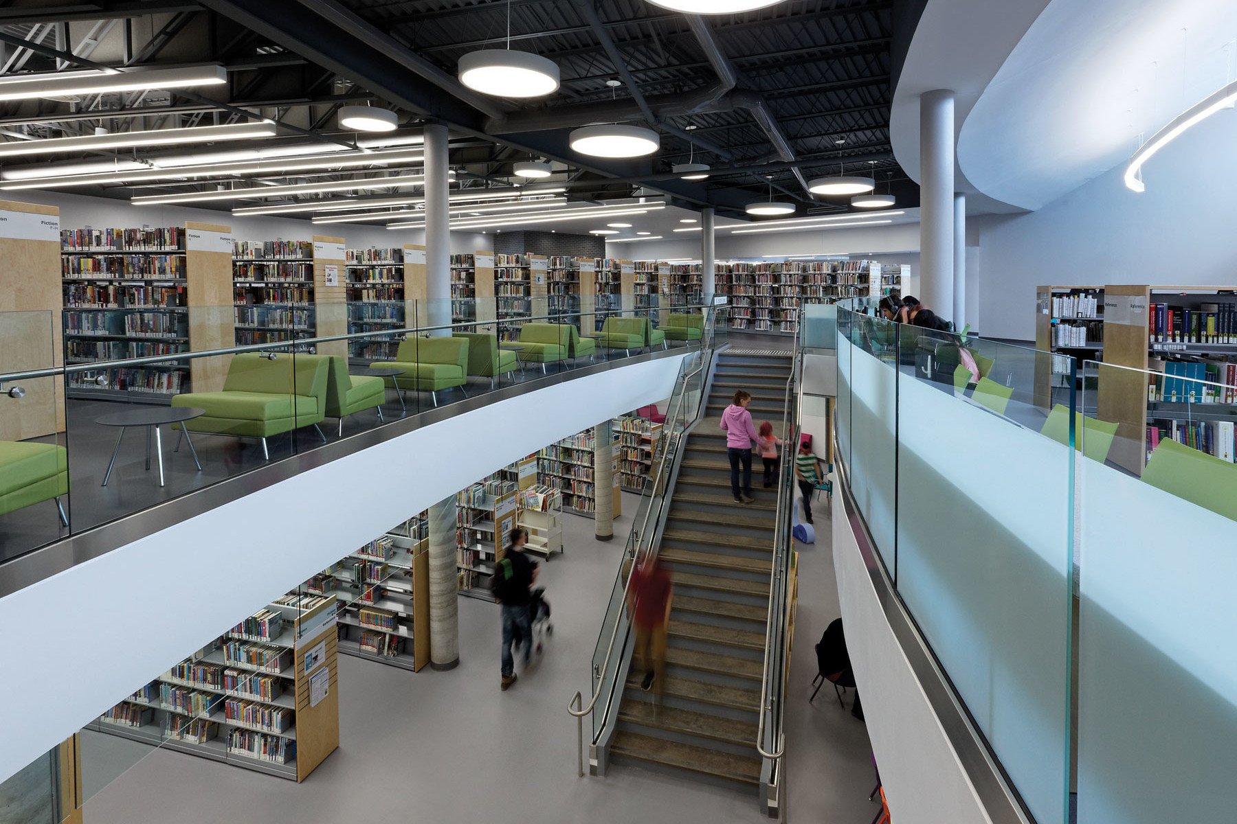 Double height space with open staircase with bookshelves on first and second storey with green lounge seating along glass guard rail