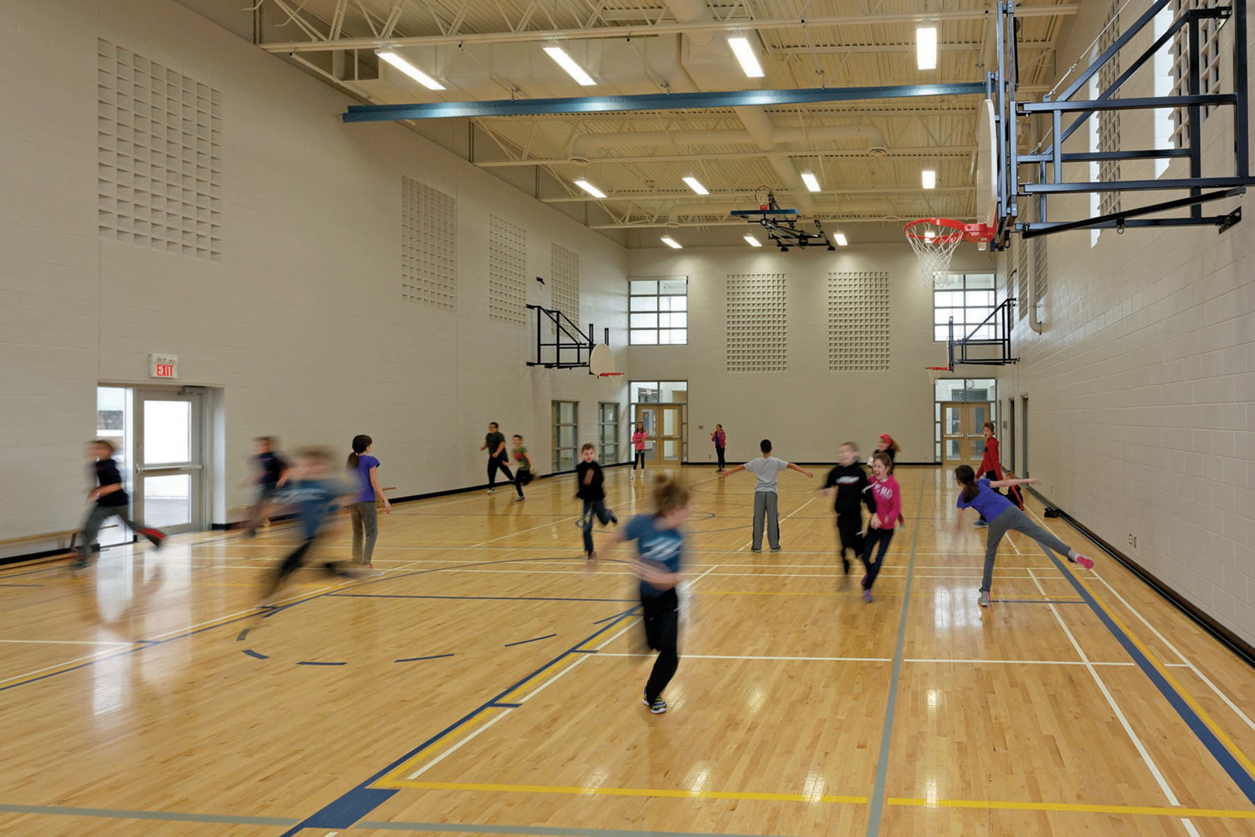 Students playing dodgeball in gymnasium with basketball nets and exposed white ceiling structure