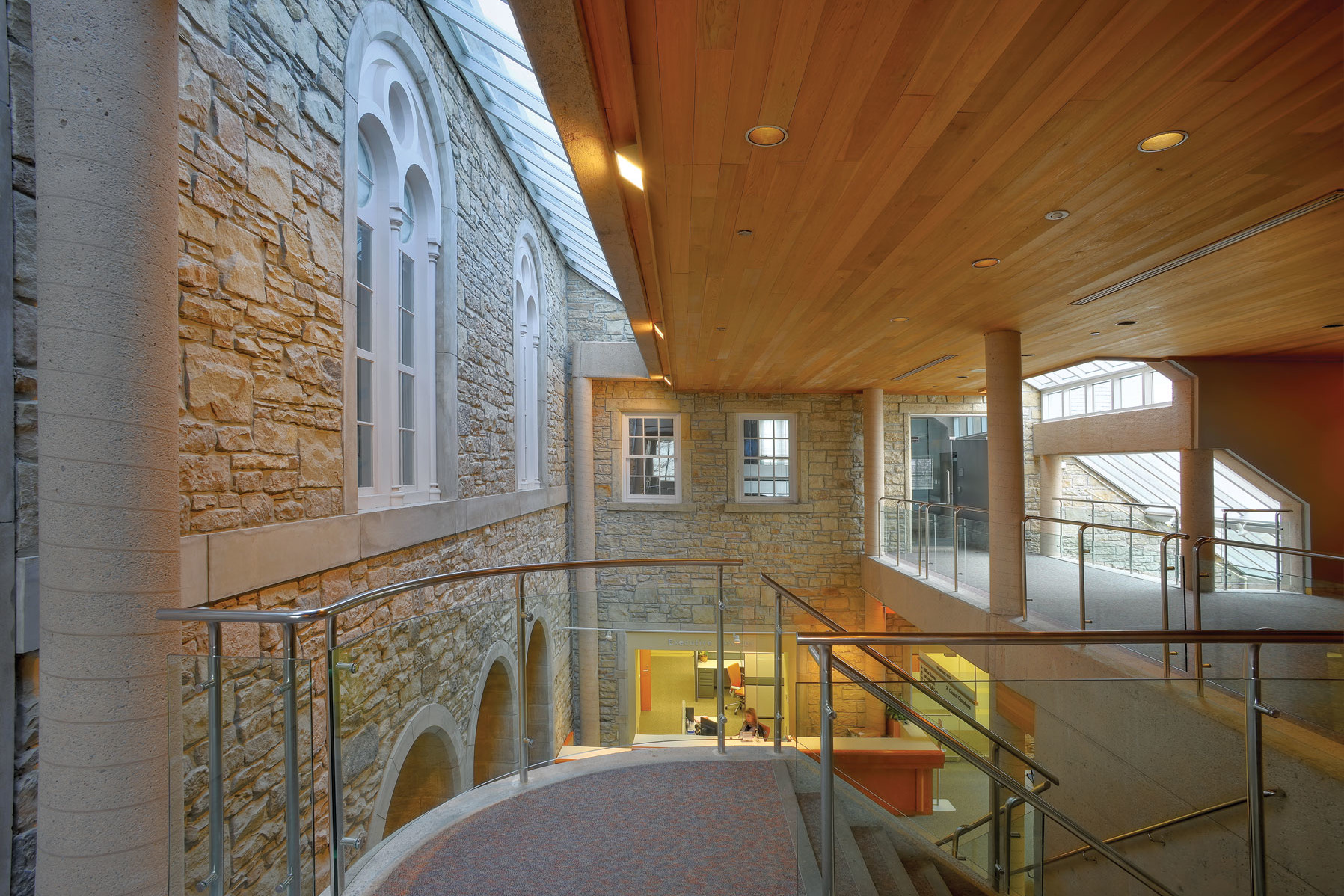 Original stone façade with arched windows connecting to addition with clerestory windows, wood plank ceiling, exposed columns and staircase