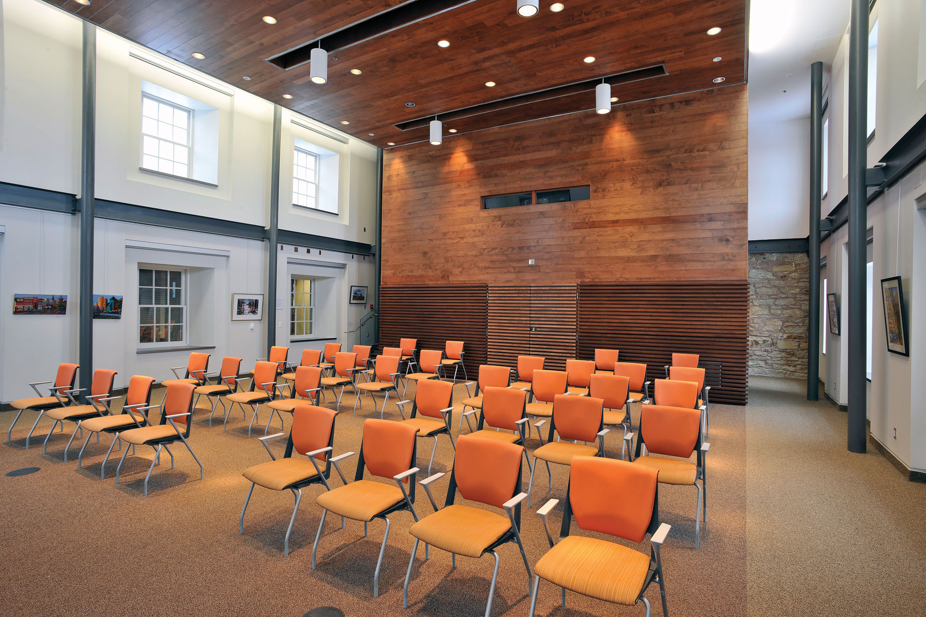 Double height public Town Hall space with windows, exposed steel beams, plank wood wall and ceiling feature, and orange chairs