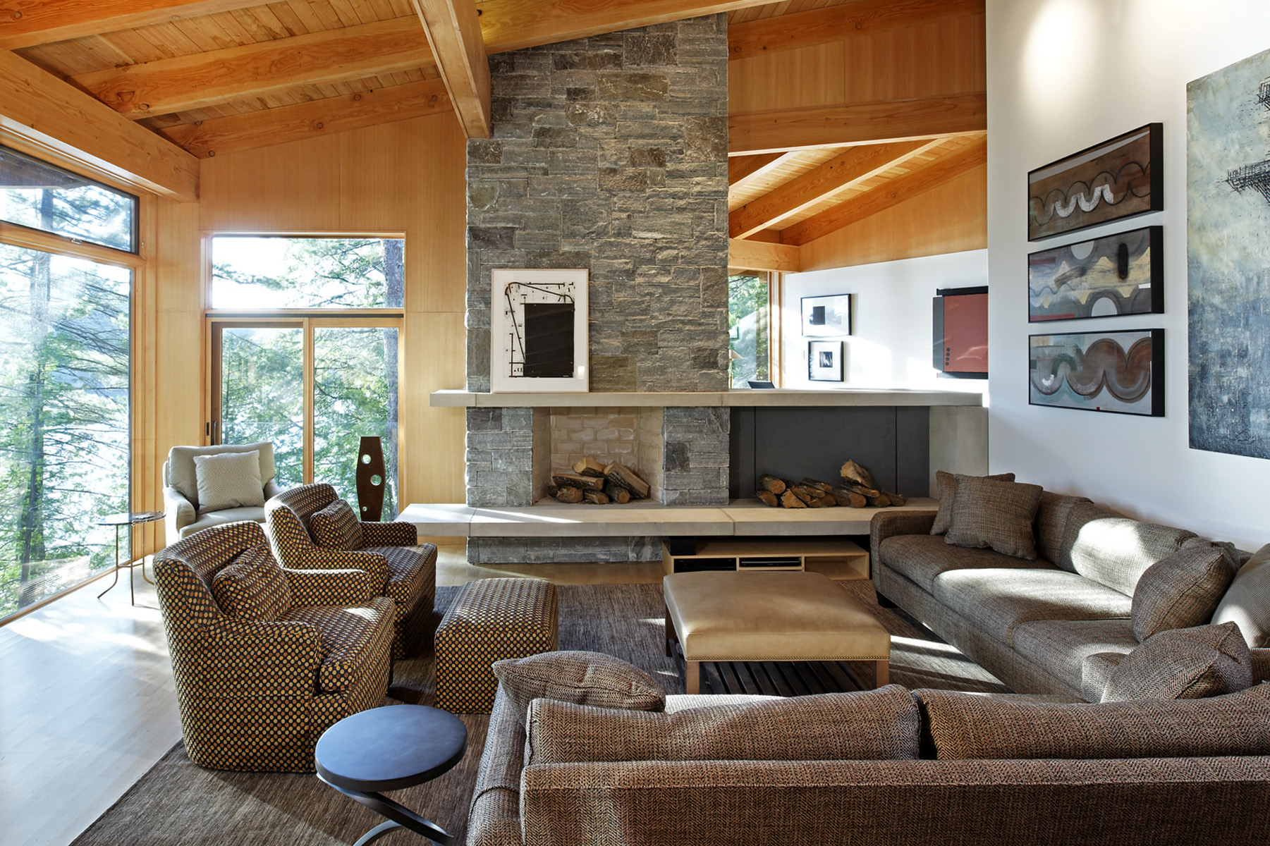 Living room with feature floor to ceiling stone fireplace and slanted wood rafter ceiling and glazed wall on left with view of trees