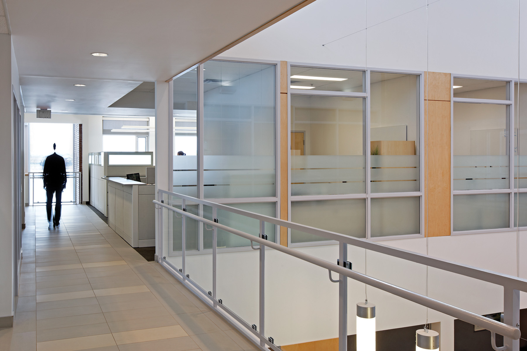 Open second storey corridor with glass partition overlooking double height front entrance atrium with workstations at end of corridor