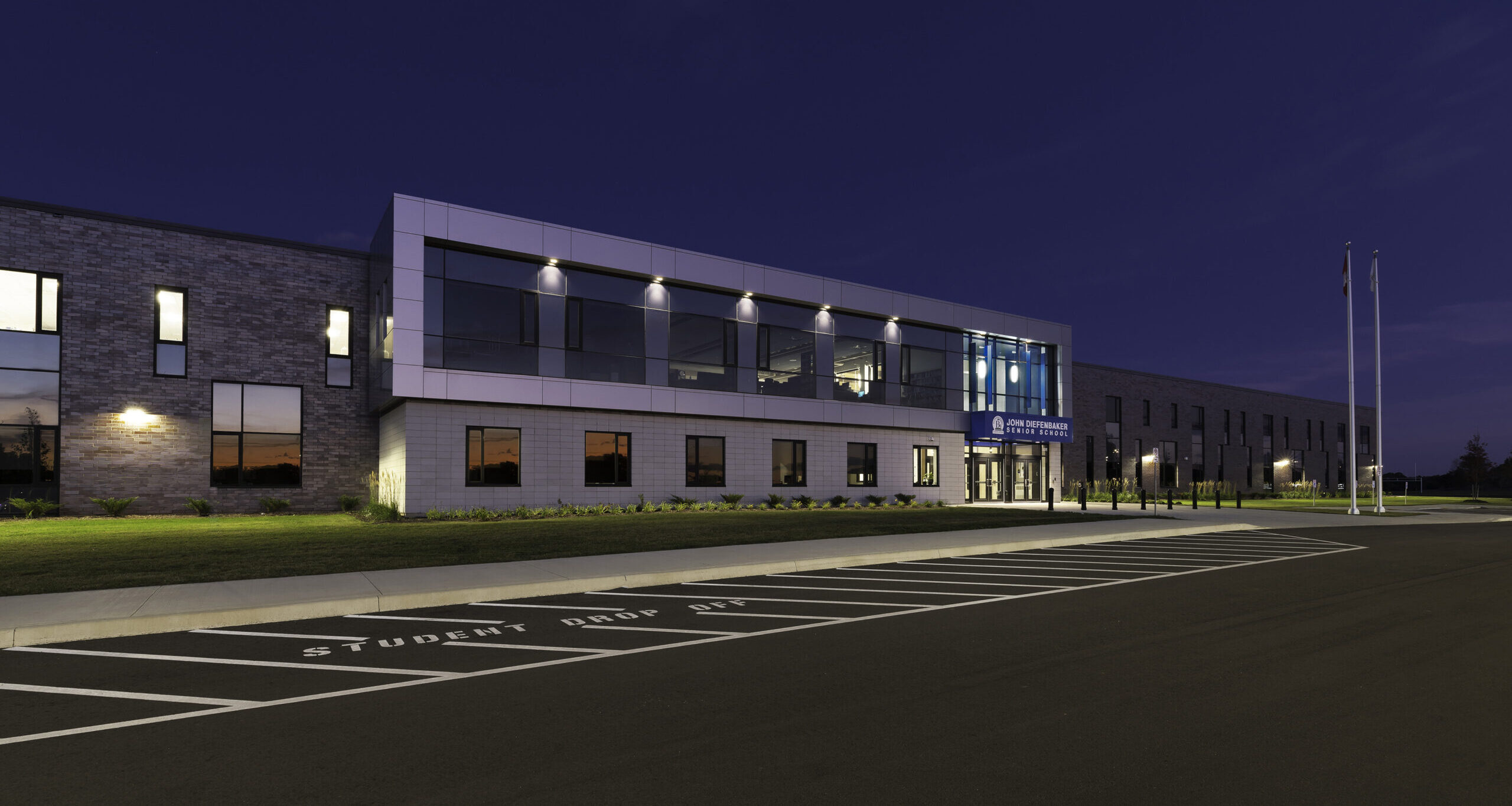 A modern school building at dusk features large windows, brick and white accents, illuminated interiors, and two flagpoles out front. The parking lot has clear markings, including a student drop-off area.