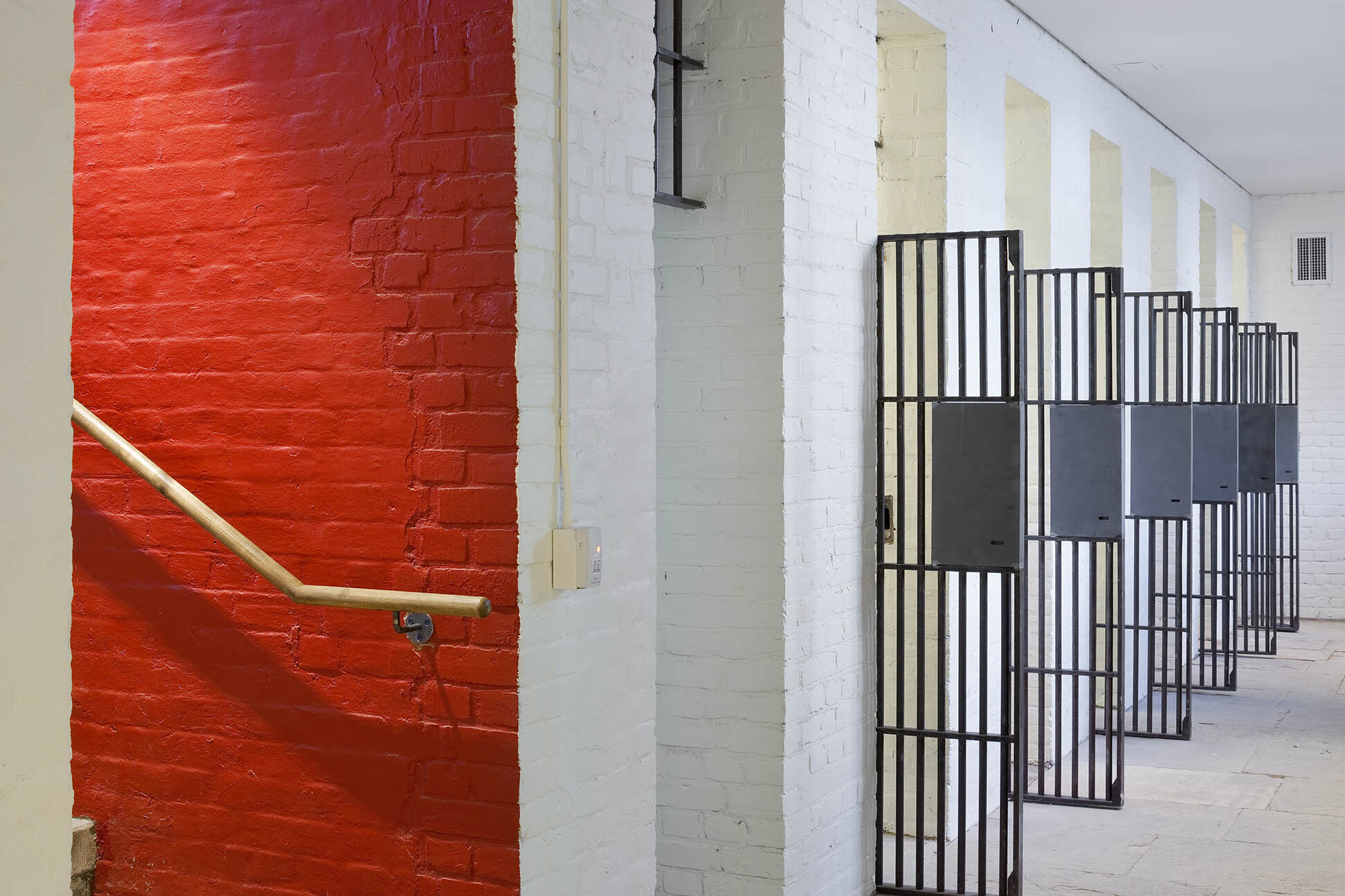 Staircase with red painted brick wall leading to white painted brick corridor with original jail cells with open barred doors