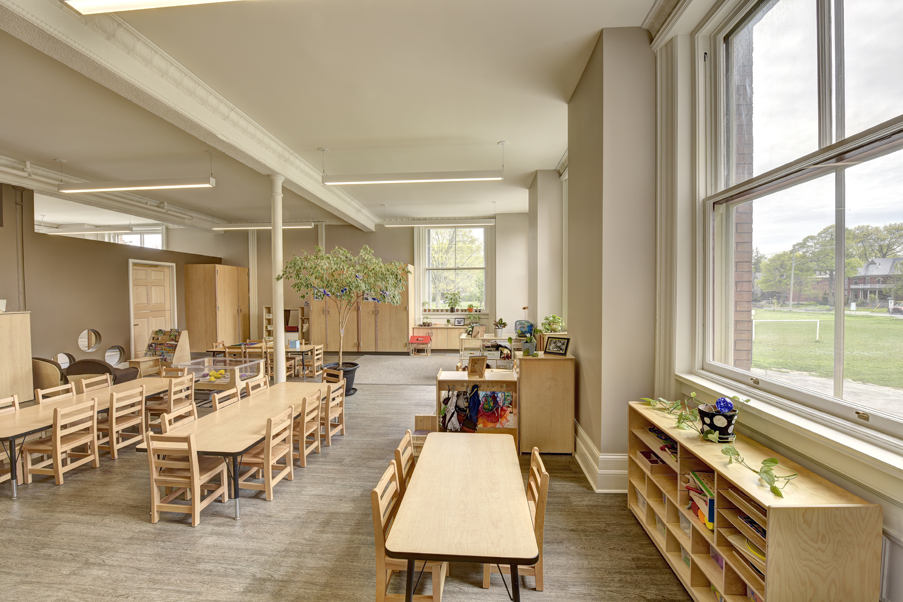 Children's classroom with long wooden tables and chairs, bookshelves, storage cupboards and large bright windows
