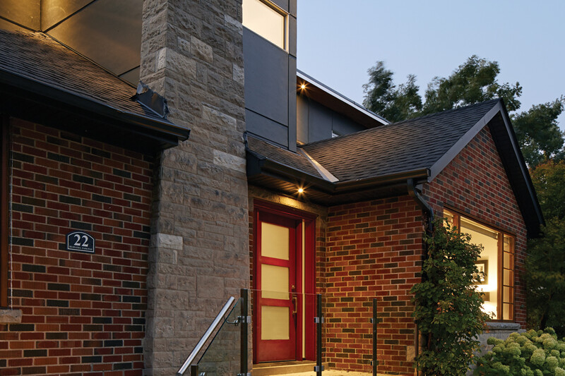 Illuminated original red brick house with second storey addition in the back and landscaped front yard at dusk