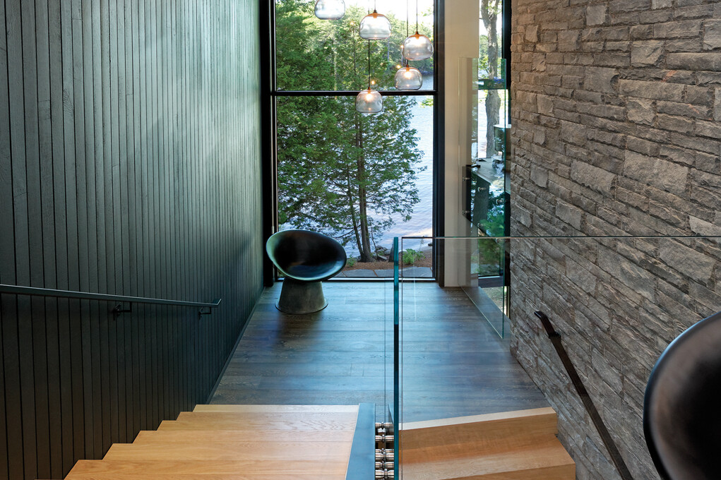 Wood stair case with glass partition with wood wall on left and stone wall on right and double height window with views of trees and lake
