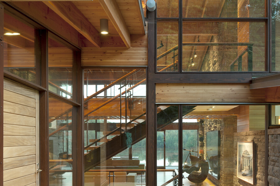 View through glazed house including living room, staircase and second storey with lake and trees on the other side of house
