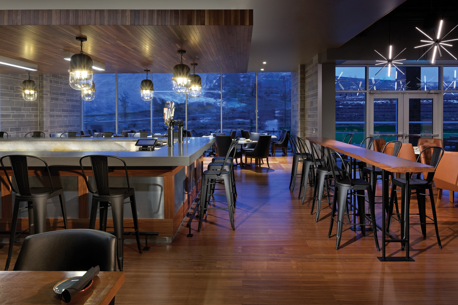 Square bar with feature lighting and black bar stools and bench and banquet seating with exterior views