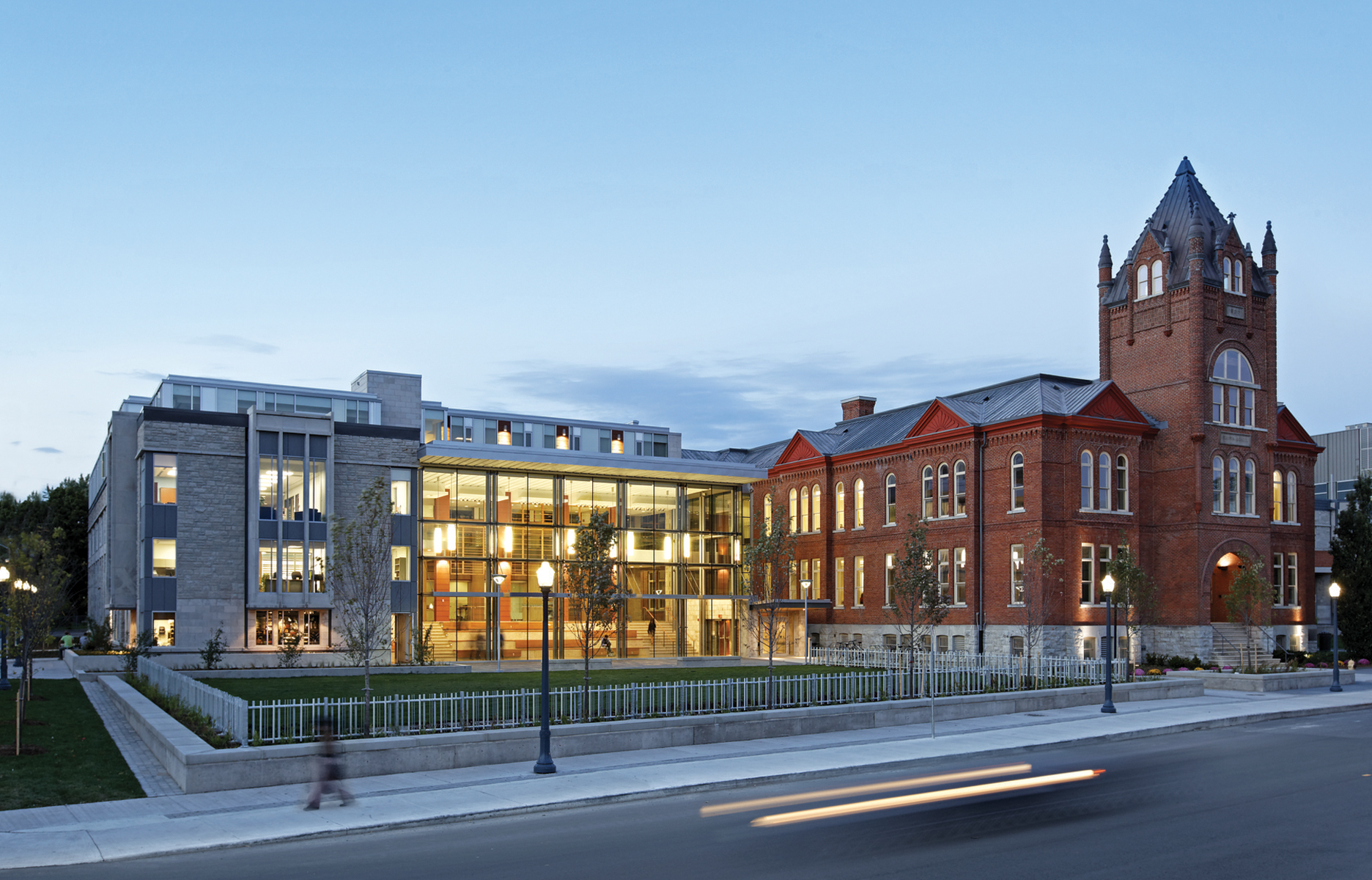 Original red brick Victoria School House with illuminated glass facade addition on left taken at dusk from road with lit lamp posts on sidewalk