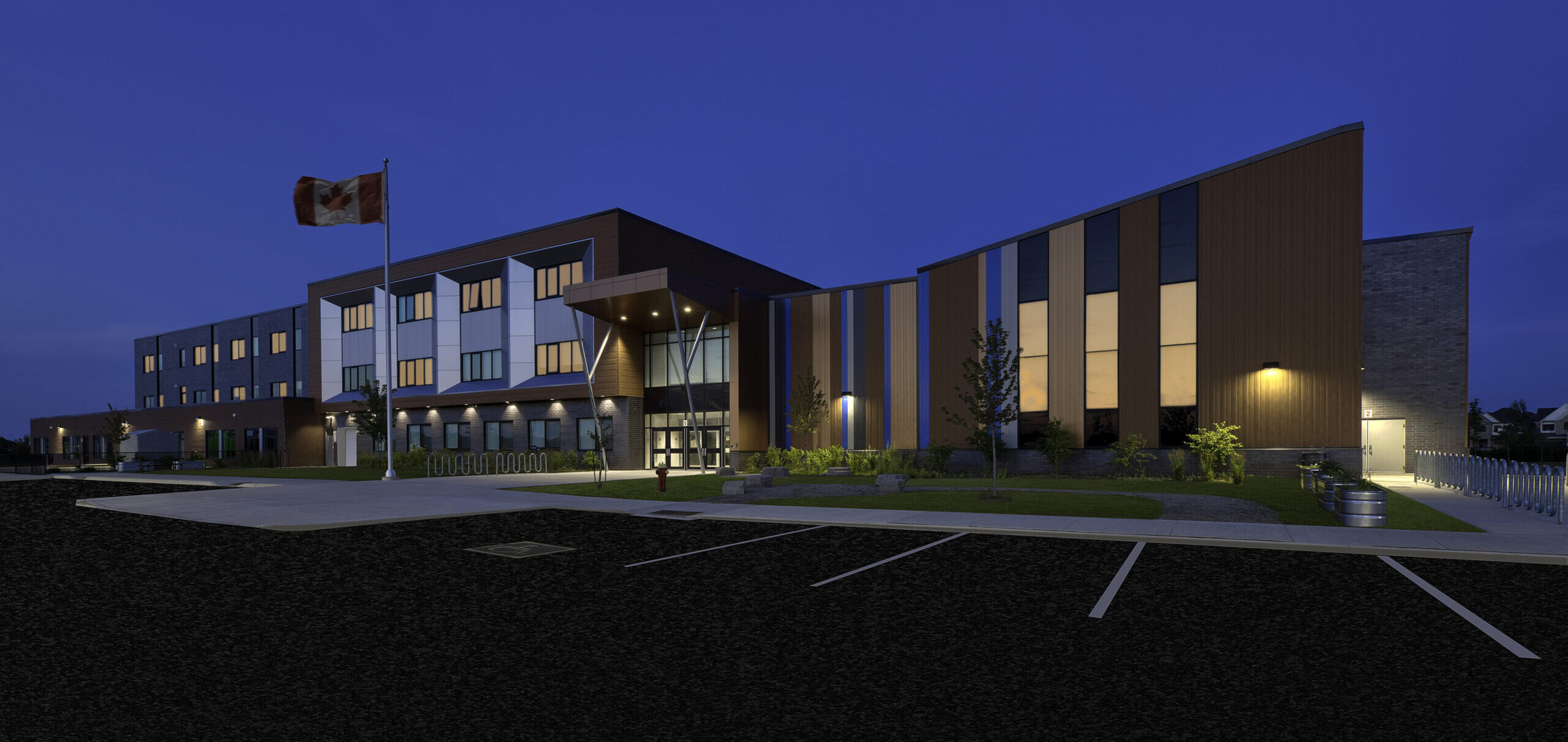 A modern, three-story building with large windows is brightly lit at dusk. A Canadian flag flies on a pole in front, and the scene includes an empty parking lot and landscaped greenery.