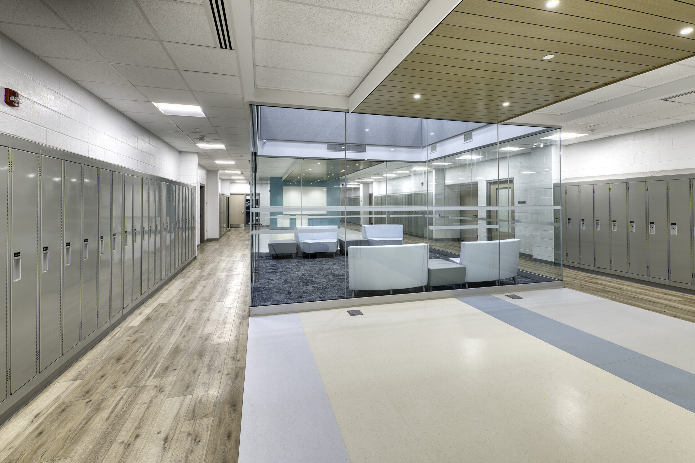 A modern school hallway with light wood floors, gray lockers on both sides, and a glass-walled lounge area in the center with white chairs and a small table. The ceiling has recessed lighting.